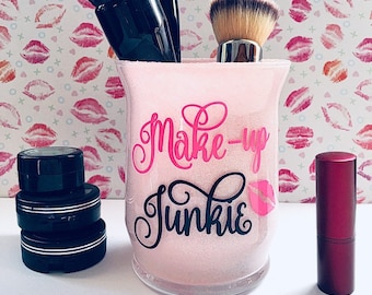 May include: Pink glitter makeup organizer with the text "Make-up Junkie" in black and pink lettering. The organizer has several makeup brushes and small containers inside. A lipstick is on the right side of the image.