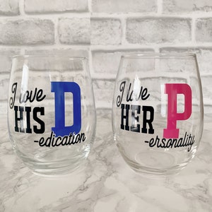 May include: Two clear wine glasses with a black and white design. The left glass says "I love his D-education" with a blue "D". The right glass says "I love her P-ersonality" with a pink "P".