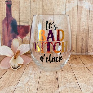 May include: A clear glass wine glass with the text "It's Bad Bitch o'clock" in black and gold lettering.