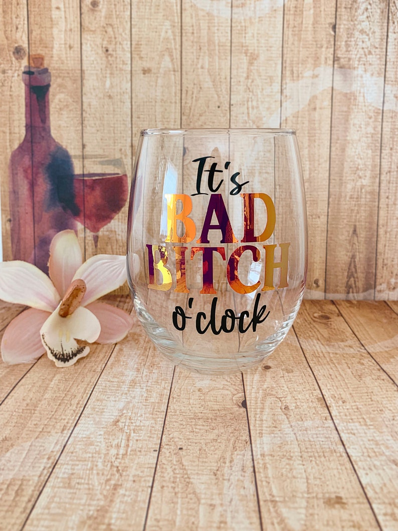 May include: A clear glass wine glass with the text "It's Bad Bitch o'clock" in black and gold lettering.