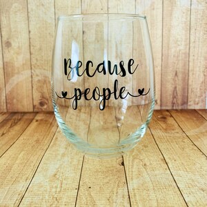 May include: A clear glass wine glass with the text "Because people" printed on the side. The glass is sitting on a wooden surface.