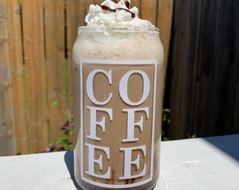 May include: A glass with a white lid and a chocolate drizzle on top. The glass is filled with a light brown liquid and has the word "COFFEE" printed on it in white.