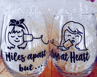 May include: Two clear glass wine glasses with black outlines of women's faces and the text "Miles apart but... close at Heart".