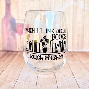May include: Clear stemless wine glass with black text that reads "When I think about books I touch my shelf." The glass has a black graphic of a bookshelf with a potted plant. A wine bottle is blurred in the background.