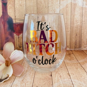 May include: A clear glass wine glass with the text "It's Bad Bitch o'clock" in gold lettering.