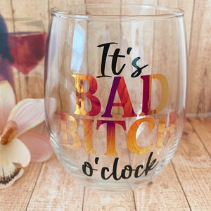 May include: A clear glass wine glass with the text "It's Bad Bitch o'clock" in black and gold foil lettering.