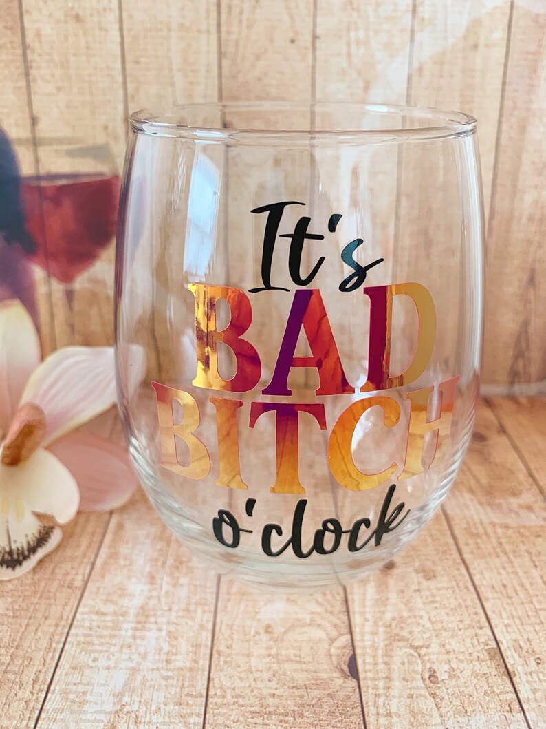 May include: A clear glass wine glass with the text "It's Bad Bitch o'clock" in black and gold foil lettering.