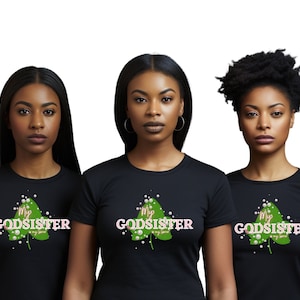 May include: Three women wearing black t-shirts with a green leaf design and the text "My Godsister" in pink and white letters.
