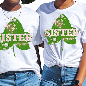 May include: Two white t-shirts with a green leaf design and the text "My Sister is my Soror" in pink letters. The shirts are worn by two people and are paired with blue jeans.