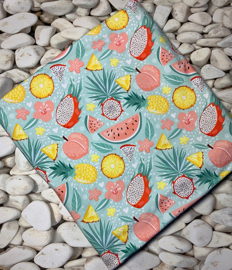 May include: A folded fabric with a tropical fruit pattern. The design features dragon fruit, watermelon, pineapple, and peaches in shades of red, yellow, and pink, set against a light blue background with floral accents.
