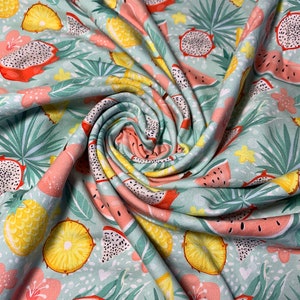 May include: A light teal fabric with a tropical fruit pattern. The design features dragon fruit, pineapples, and watermelon slices, along with pink flowers and green leaves. The fabric is soft and flowing, with a slight sheen.