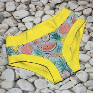 May include: A pair of underwear with a vibrant tropical fruit pattern, including watermelon, peaches, and dragon fruit, set against a light blue background. The edges are trimmed with bright yellow fabric.