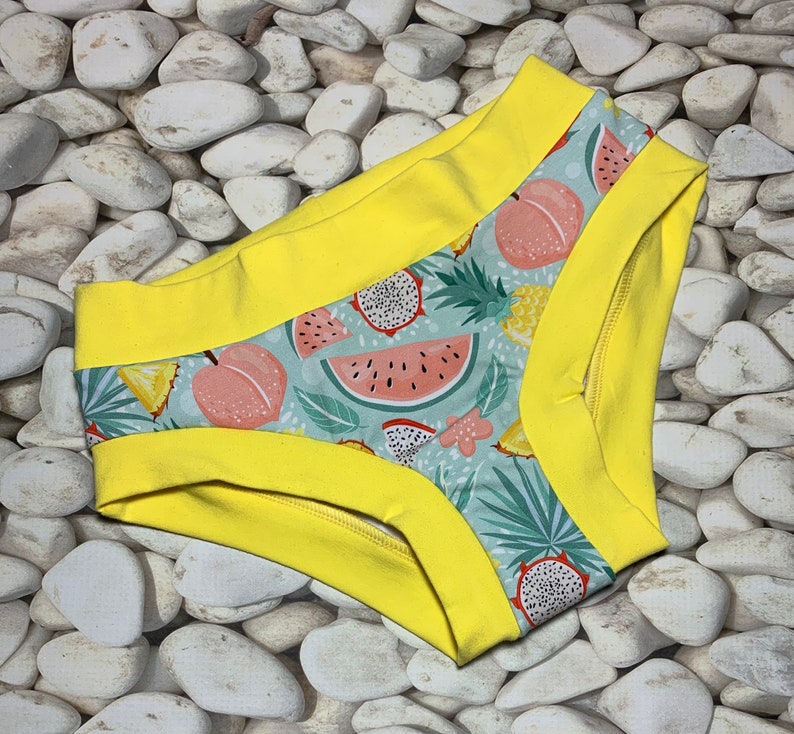 May include: A pair of underwear with a vibrant tropical fruit pattern, including watermelon, peaches, and dragon fruit, set against a light blue background. The edges are trimmed with bright yellow fabric.