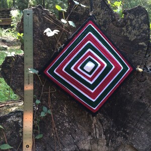 May include: A red, white, and green yarn dreamcatcher with a diamond shape hangs from a tree branch. The dreamcatcher is made with multiple layers of yarn and has a white center.
