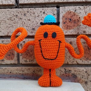 May include: A crocheted orange Mr. Men character with a blue hat and a black smile. The character has long orange arms and is standing on a white surface.