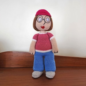 Könnte beinhalten: Eine gehäkelte Puppe mit braunen Haaren, die ein rotes Shirt, eine blaue Hose und eine rosa Mütze trägt. Die Puppe hat schwarze Augen und einen lächelnden Mund.