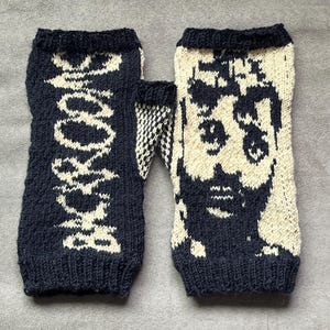 May include: A pair of fingerless gloves featuring a black and cream knit pattern. One glove displays the text "SHROOMS" in a vertical orientation, while the other glove features a detailed graphic portrait of a face with large eyes.