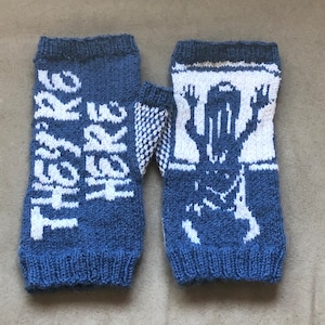 May include: A pair of blue knitted fingerless gloves with white knit designs. One glove has the text "They're Here" and the other has a stylized figure.