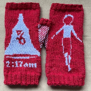 May include: Red knitted fingerless gloves with light blue graphics. One glove features a mountain design with the text "2:17 am". The other glove has a stick figure design. The gloves are made of a thick, textured yarn.