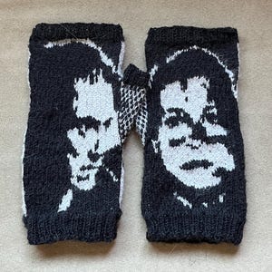 May include: A pair of black and white fingerless gloves featuring a knitted portrait design on the front of each piece. The gloves have a ribbed cuff at the top and bottom, with a contrasting black and white pattern visible on the thumb gusset.