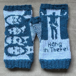 May include: A pair of knitted fingerless gloves in teal and white. One glove features a design with the words "CRY HOT" and fish shapes. The other glove has a silhouette of a person and the words "Hang In There!"