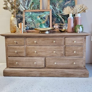 May include: A wooden dresser with nine drawers and brass knobs. The dresser is decorated with three framed abstract paintings with green and gold swirls, two vases with dried palm leaves, and a gold metal tray.