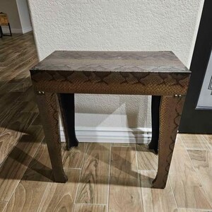 Puede incluir: Una mesa auxiliar rectangular con un acabado de patrón de piel de serpiente en tonos marrón y negro. La mesa tiene cuatro patas con una ligera curva hacia afuera y está decorada con pequeños remaches metálicos circulares en la parte superior.
