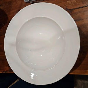 May include: A round, white ceramic pasta bowl featuring a wide, flat rim and a deep, concave center. The bowl has a smooth, glossy finish and a clean, minimalist design, providing a neutral surface for serving various food dishes.