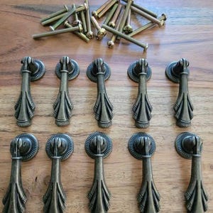 May include: A set of ten antique brass-toned metal drop pulls for furniture, each featuring a circular mounting plate and a flared, textured handle. The collection includes a matching set of ten long metal screws for installation.