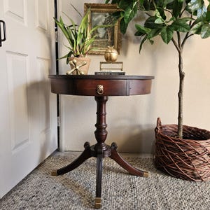 Puede incluir: Una mesa auxiliar redonda de madera oscura con un solo cajón y un tirador con cabeza de león. La mesa tiene tres patas con detalles dorados y se encuentra sobre una alfombra gris. Una planta en maceta y un globo terráqueo dorado están sobre la mesa.