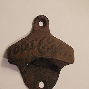 May include: A vintage cast iron Coca-Cola bottle opener with a brown patina. The opener has a hole at the top for hanging and a large opening for opening bottles.