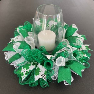May include: A green and white wreath with shamrock print fabric and green glitter ribbon. A clear glass votive holder with a white candle sits in the center of the wreath.