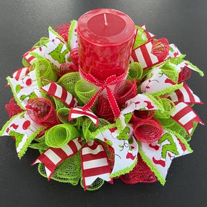 May include: A Christmas centerpiece featuring a red pillar candle surrounded by red and green mesh, white ribbon with red polka dots, and red and white striped ribbon. The ribbon has a Grinch-themed print.