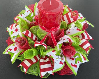 May include: A Christmas centerpiece featuring a red pillar candle surrounded by red and green mesh, white ribbon with red polka dots, and red and white striped ribbon. The ribbon has a Grinch-themed print.