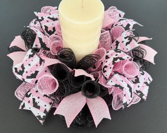 Puede incluir: Una vela de pilar color crema se encuentra en el centro de una corona decorativa. La corona presenta cintas de malla rosa y negra, con patrones de murciélagos y rayas. El diseño general es un portavelas temático de Halloween.