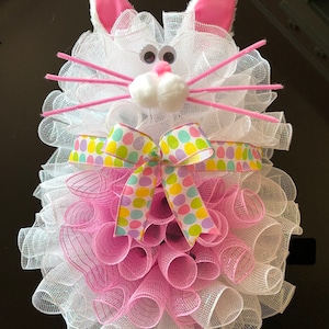 May include: A white bunny-shaped Easter wreath with pink ears, a white pom-pom nose, and pink and white mesh. The wreath is decorated with a pink and green polka dot ribbon.