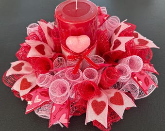 Può includere: Un centrotavola per San Valentino con una candela rossa a forma di pilastro sormontata da un cuore rosa, circondata da tulle rosa e rosso e fiocchi di nastro con cuori glitterati.