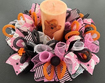 Puede incluir: Un centro de mesa con velas de Halloween. La vela de color melocotón tiene una decoración de oso brillante. La base es una corona de cintas rosas, negras y naranjas, con acentos de fantasmas y calabazas. Un artículo festivo y decorativo.