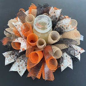 May include: A Halloween-themed wreath with orange, black, and brown mesh, accented with burlap and ribbon. A clear glass jar candle sits in the center of the wreath.