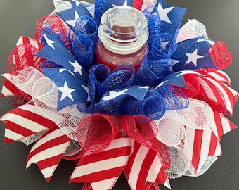 May include: A patriotic candle holder featuring a red candle in a glass jar, surrounded by red, white, and blue ribbons and mesh. The ribbons are decorated with white stars and red and white stripes, creating a festive, decorative piece.