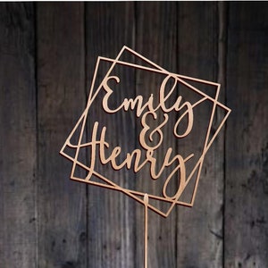 May include: A wooden cake topper with the names "Emily & Henry" in a geometric frame. The topper is on a wooden background.