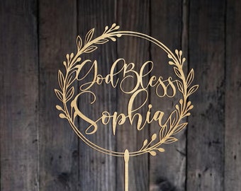 Op de afbeelding: Een houten taarttopper met een rond kransontwerp. De krans is gemaakt van bladeren en heeft de woorden "God Bless Sophia" erin geschreven. De topper zit op een houten stok.