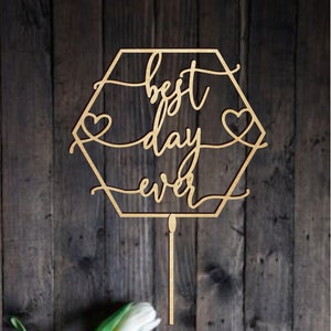 Può includere: Topper per torta in legno con una cornice esagonale e il testo "best day ever" in corsivo. Due piccoli cuori sono inclusi su entrambi i lati del testo. Il topper è su uno sfondo di legno con tulipani bianchi.