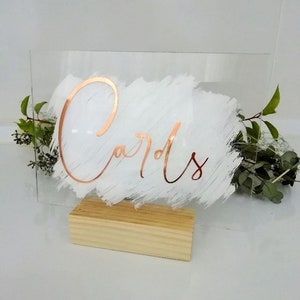 May include: A clear acrylic sign with a white brushstroke background and the word "Cards" written in copper foil lettering. The sign is on a small wooden stand.