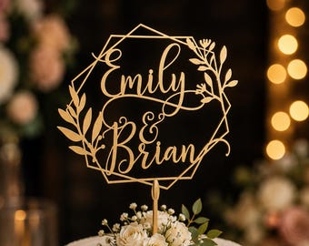 Puede incluir: Un adorno para pastel de color dorado que presenta un marco hexagonal geométrico con los nombres Emily & Brian en letra cursiva, acentuado con recortes de hojas y flores, colocado sobre un pastel glaseado en blanco decorado con rosas blancas.