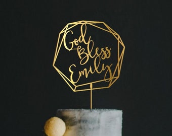 Op de afbeelding: Gouden geometrische taarttopper met de tekst "God Bless Emily" op een grijze tweelaagse taart.