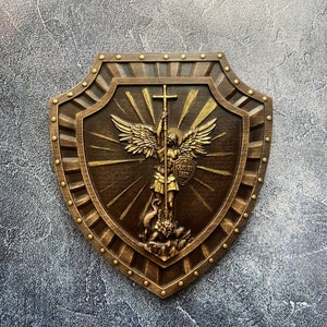 May include: A brown wooden shield with a gold-painted design of an angel holding a cross and a sword. The angel is standing on a dragon. The shield has a gold border with decorative studs. The text "QUIS UT DEUS" is written on the shield.