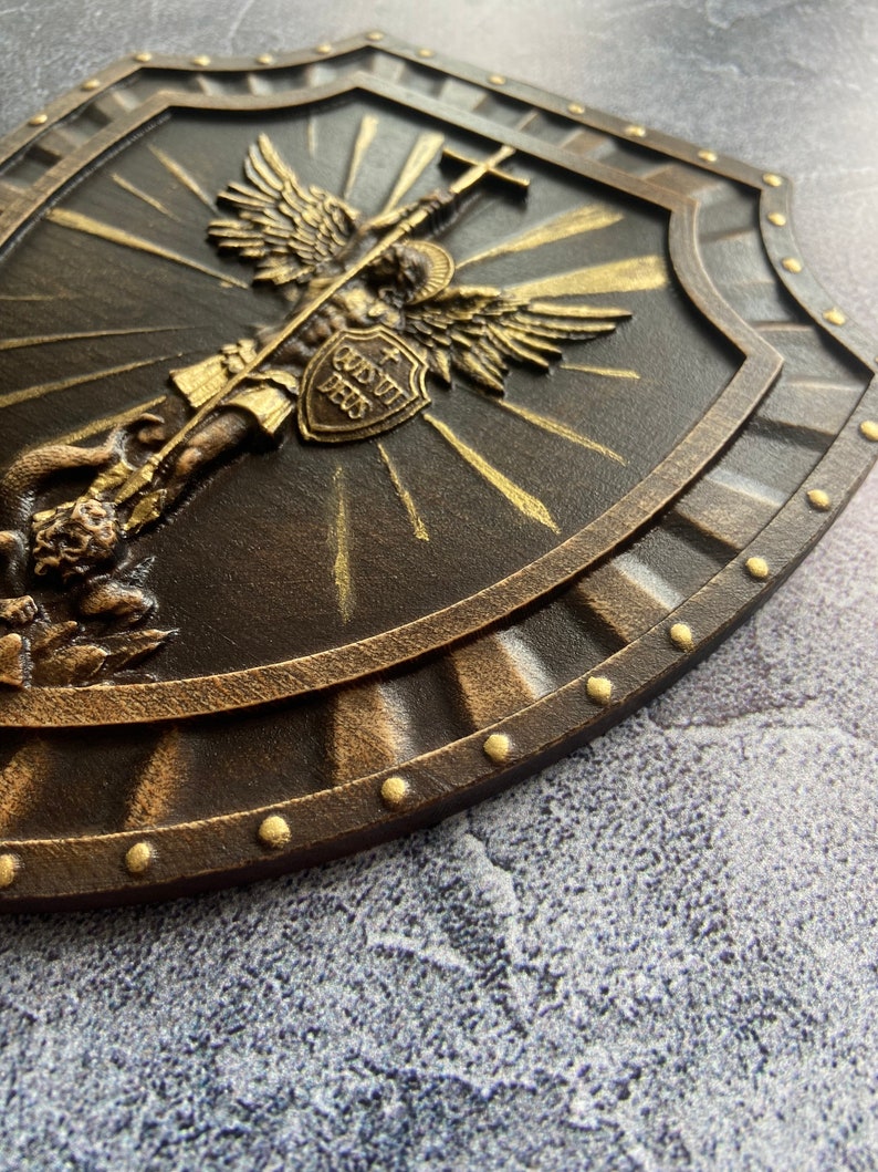 May include: A brown wooden shield with a gold-colored angel holding a sword and a cross. The angel is surrounded by a golden halo and rays of light. The shield has a gold-colored border and is decorated with gold-colored studs. The shield has the words "Custo Dei" written on it.