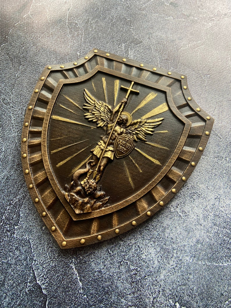 May include: A brown wooden shield with a gold-painted design of an angel holding a cross and a sword. The angel is standing on a dragon. The shield has a gold-painted border and is decorated with gold-painted studs. The shield has the text "In Quo Confido Deus" on it.