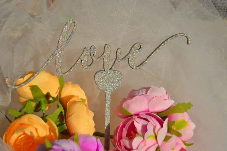 Puede incluir: Un letrero de 'Love' de purpurina plateada con un coraz&oacute;n en el centro, rodeado de un ramo de flores rosas y naranjas.